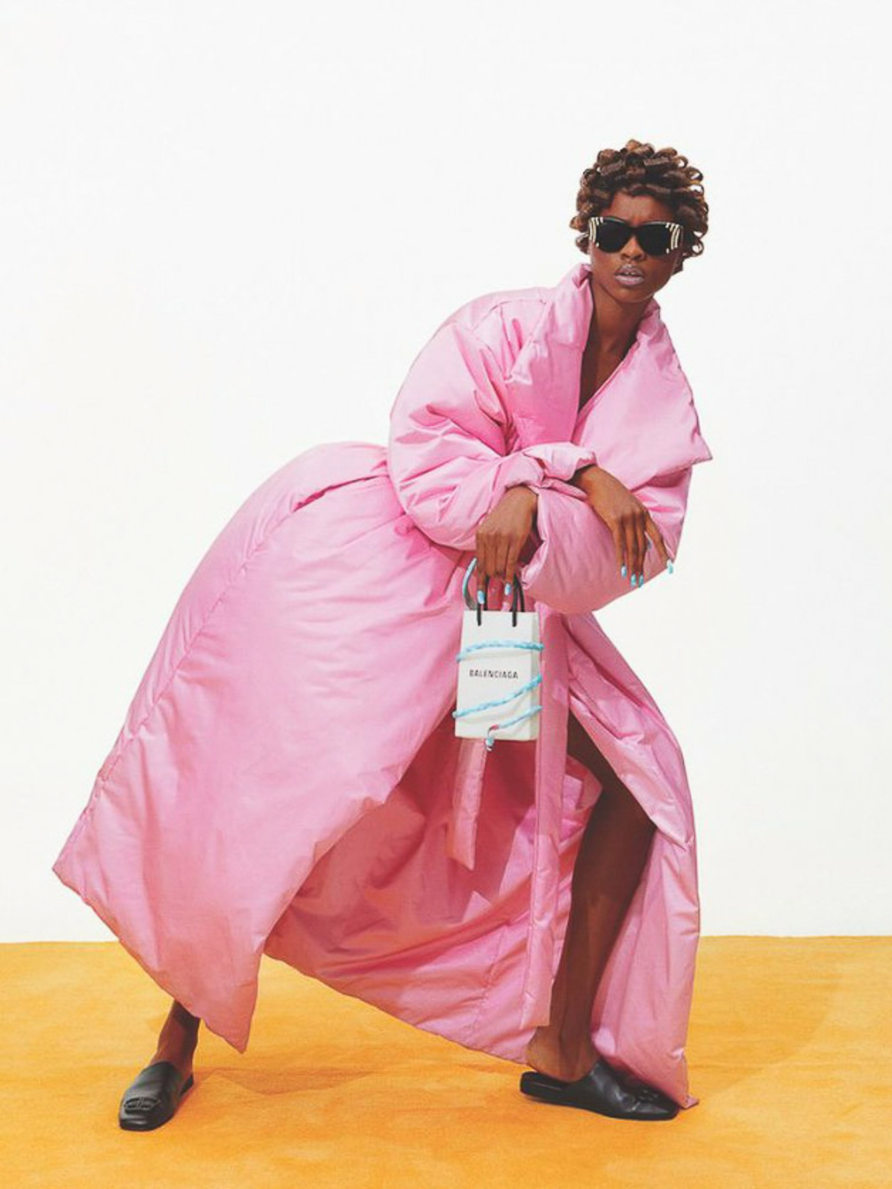 A model wearing a voluminous pink coat and black sunglasses poses with a Balenciaga handbag against a yellow and white backdrop.