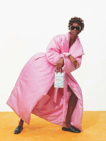 A model wearing a voluminous pink coat and black sunglasses poses with a Balenciaga handbag against a yellow and white backdrop.