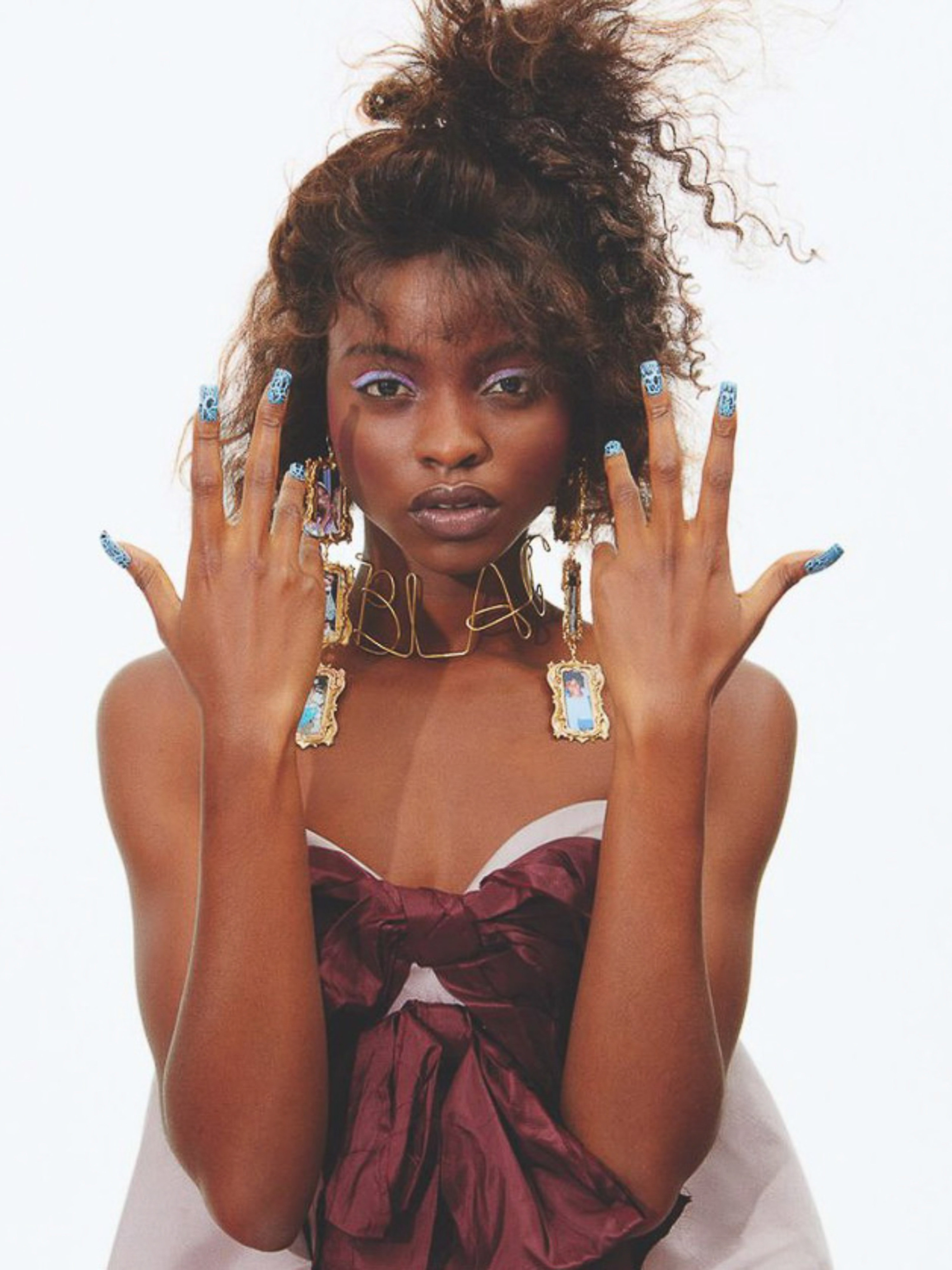 Portrait of a Black woman with elaborate nail art, gold necklace spelling 'BLA,' and a burgundy ribbon against a plain white background.