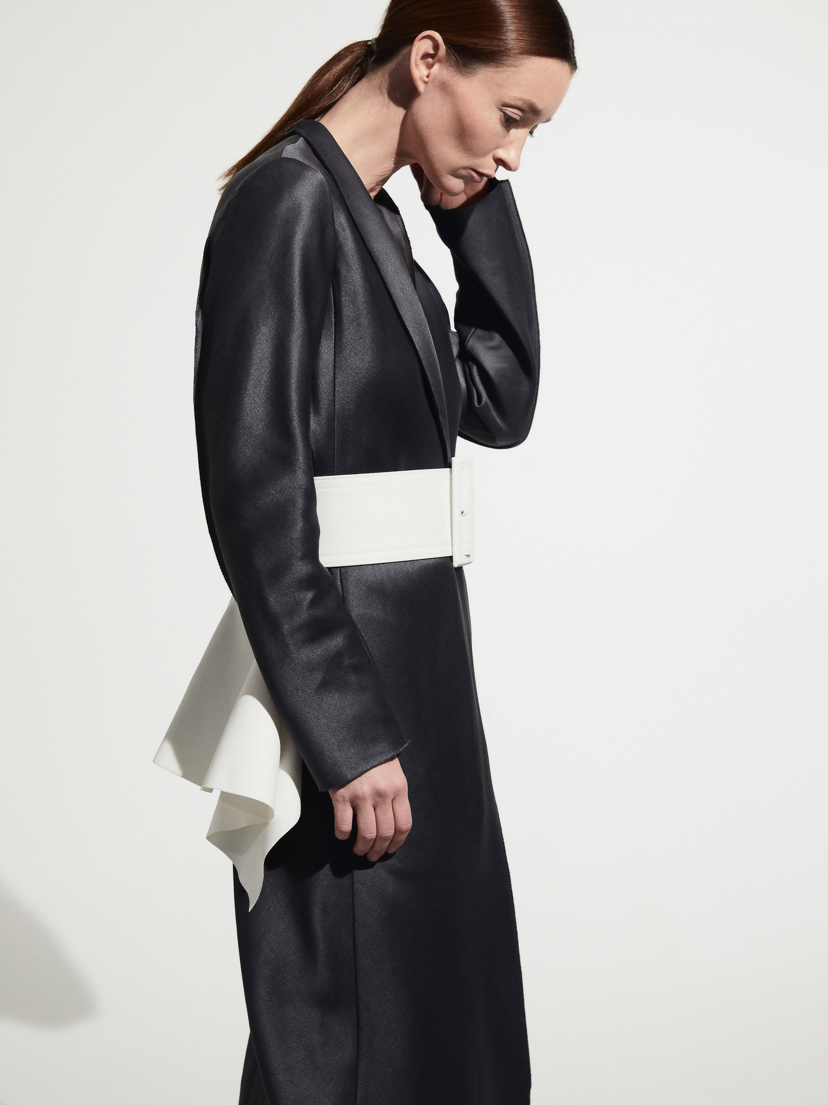 Elegant woman in a dark grey dress with a dramatic white sash, side view against a white backdrop. Fashion photography, minimal style.