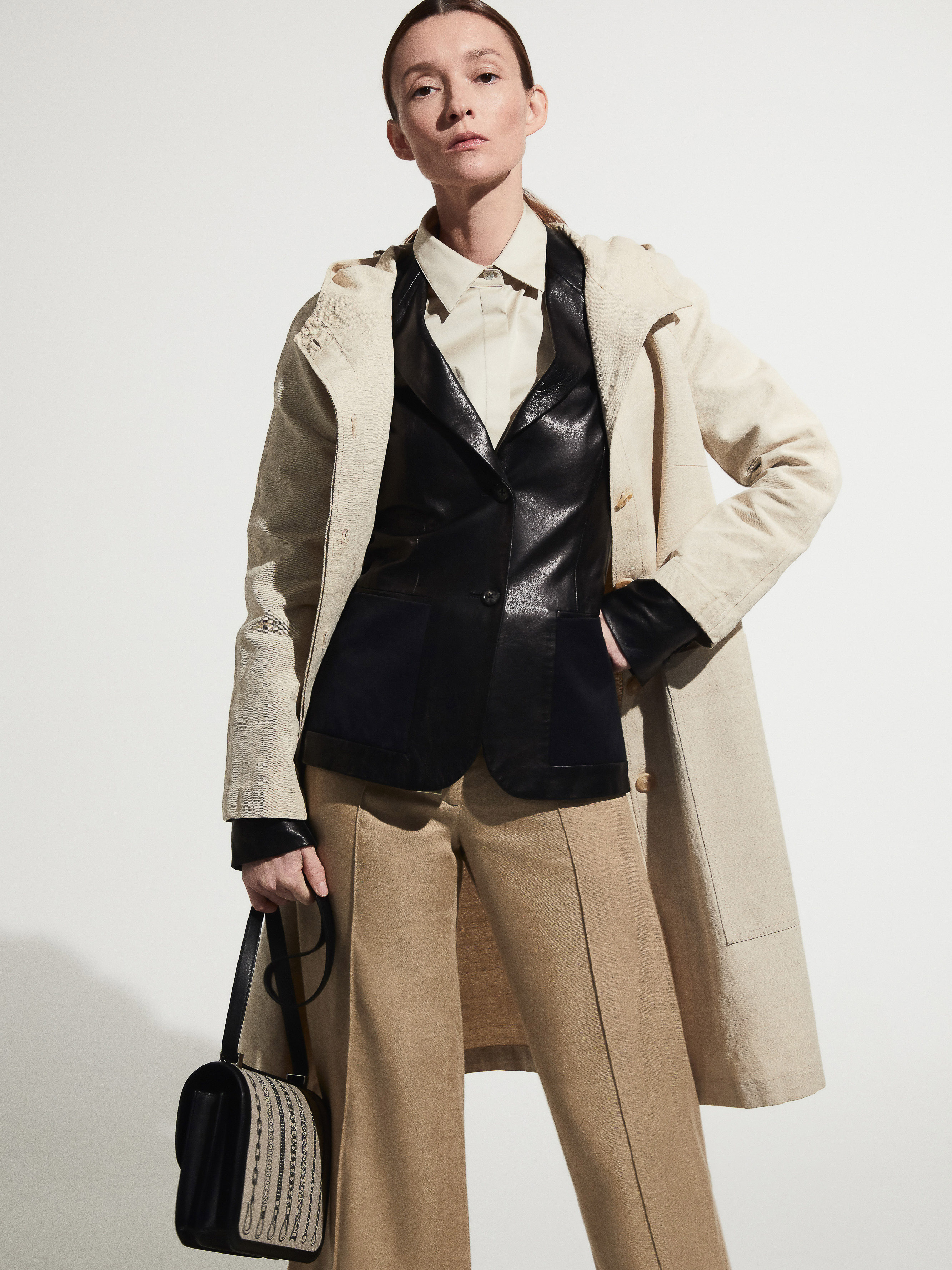 A woman stands in a studio wearing a tan coat, black vest, khaki pants, and black bag, posing against a white backdrop.