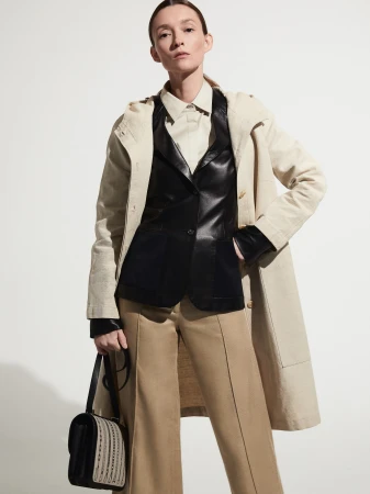 A woman stands in a studio wearing a tan coat, black vest, khaki pants, and black bag, posing against a white backdrop.