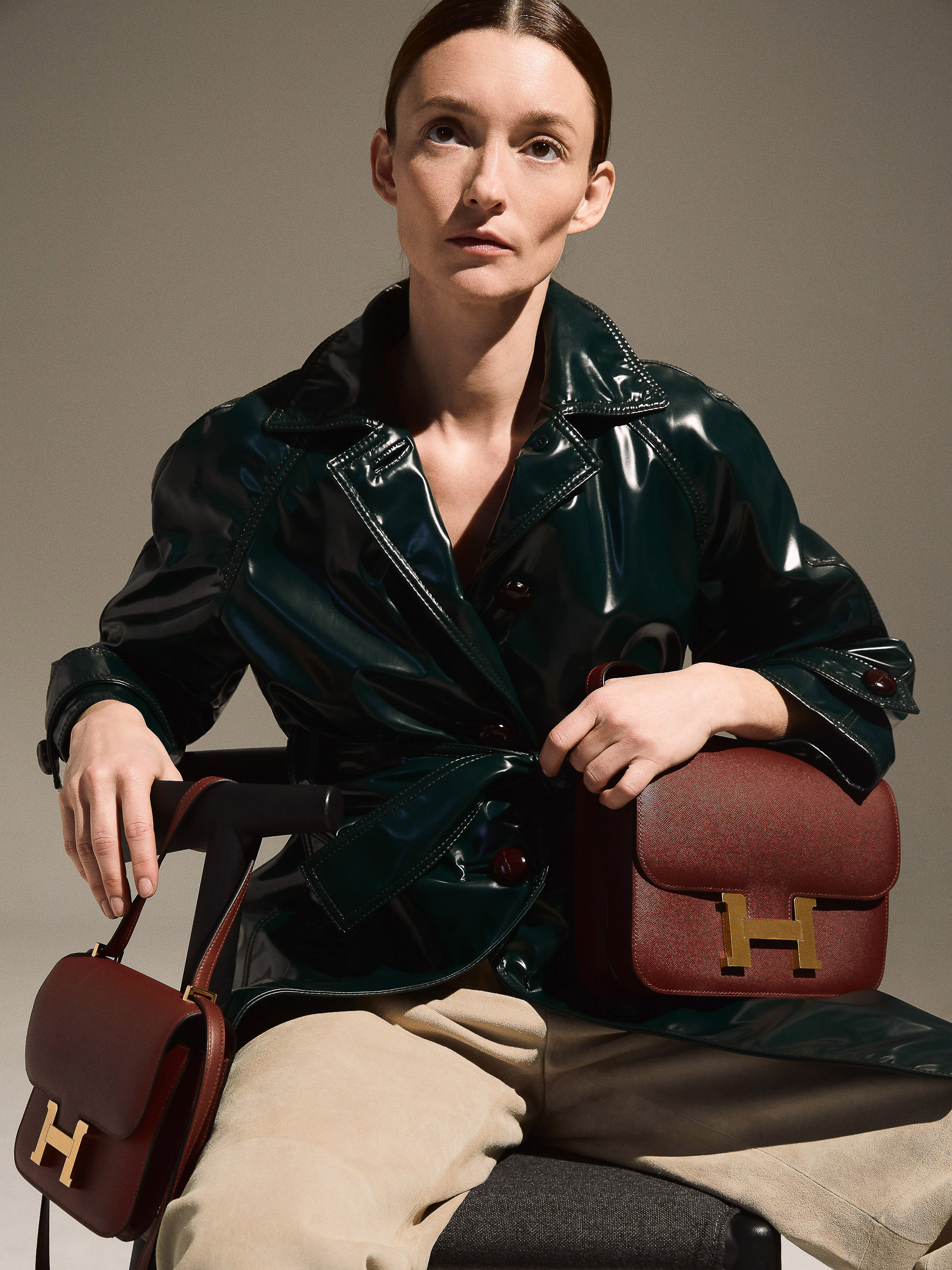 A woman poses with two brown leather handbags featuring gold hardware in a studio setting, wearing a shiny green trench coat and beige pants.