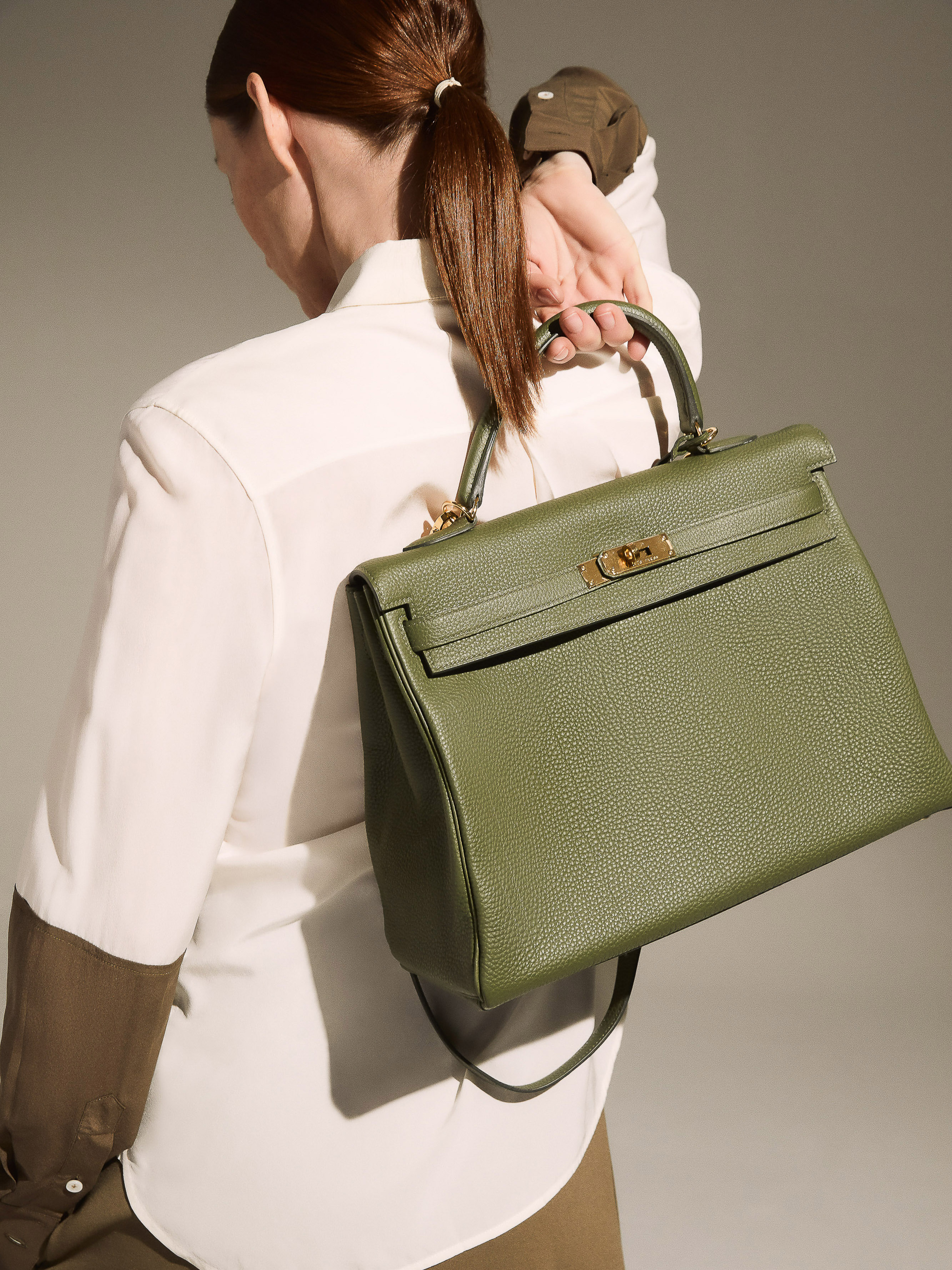 Olive-green textured leather Kelly bag with gold hardware. Woman in white shirt holding designer handbag. Studio shot.