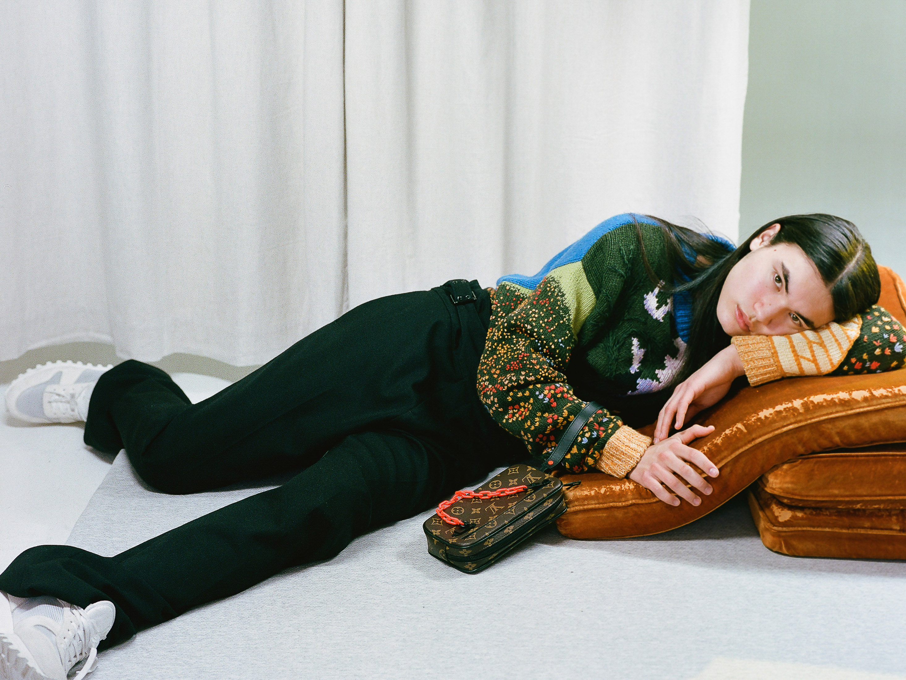 A young woman in a multicolored knit sweater reclines on a brown pillow with a designer handbag, sneakers, and curtains in background.