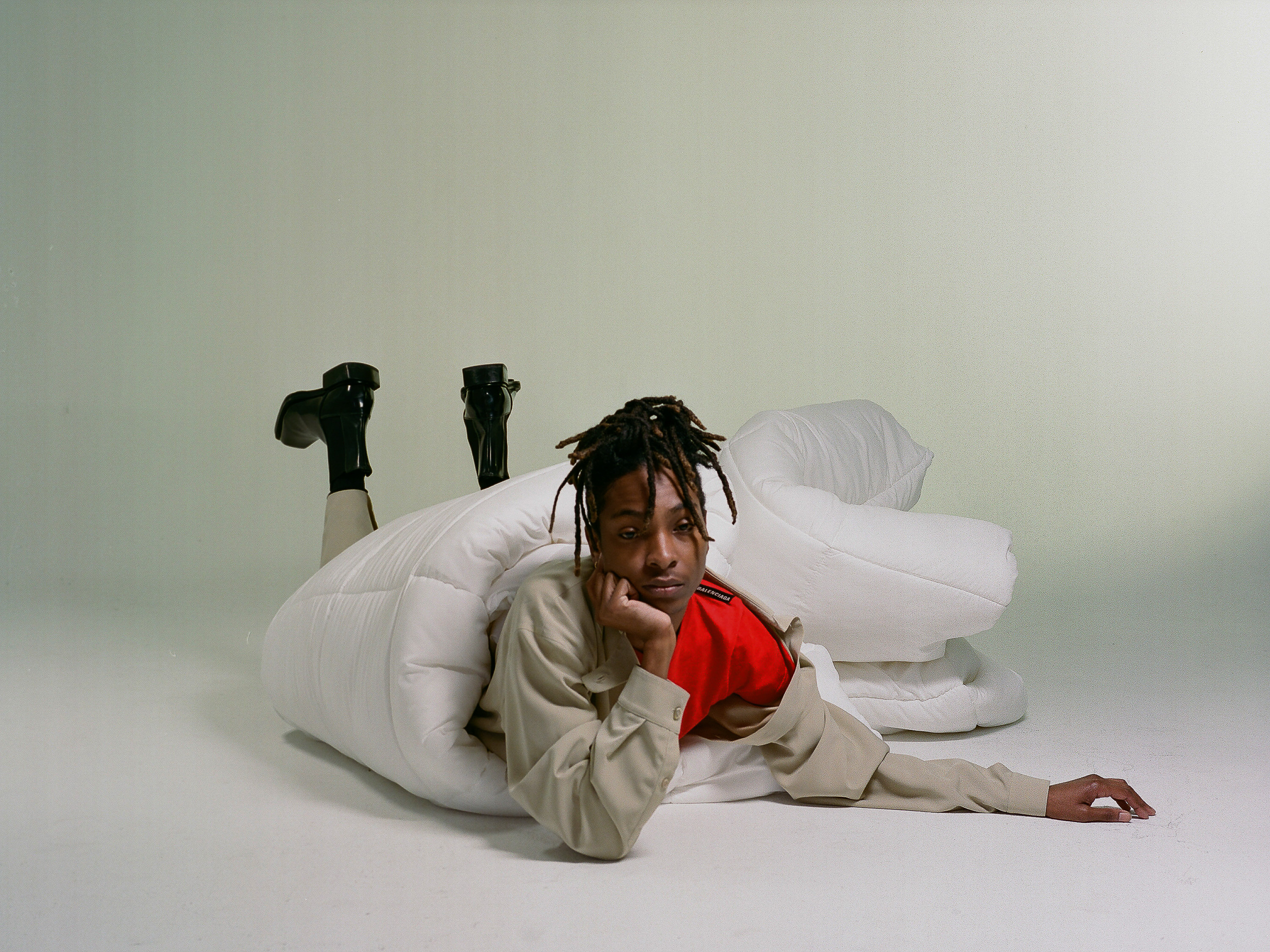 Person in an oversized white puffy comforter with black heeled boots against a light grey backdrop in a studio fashion shoot.