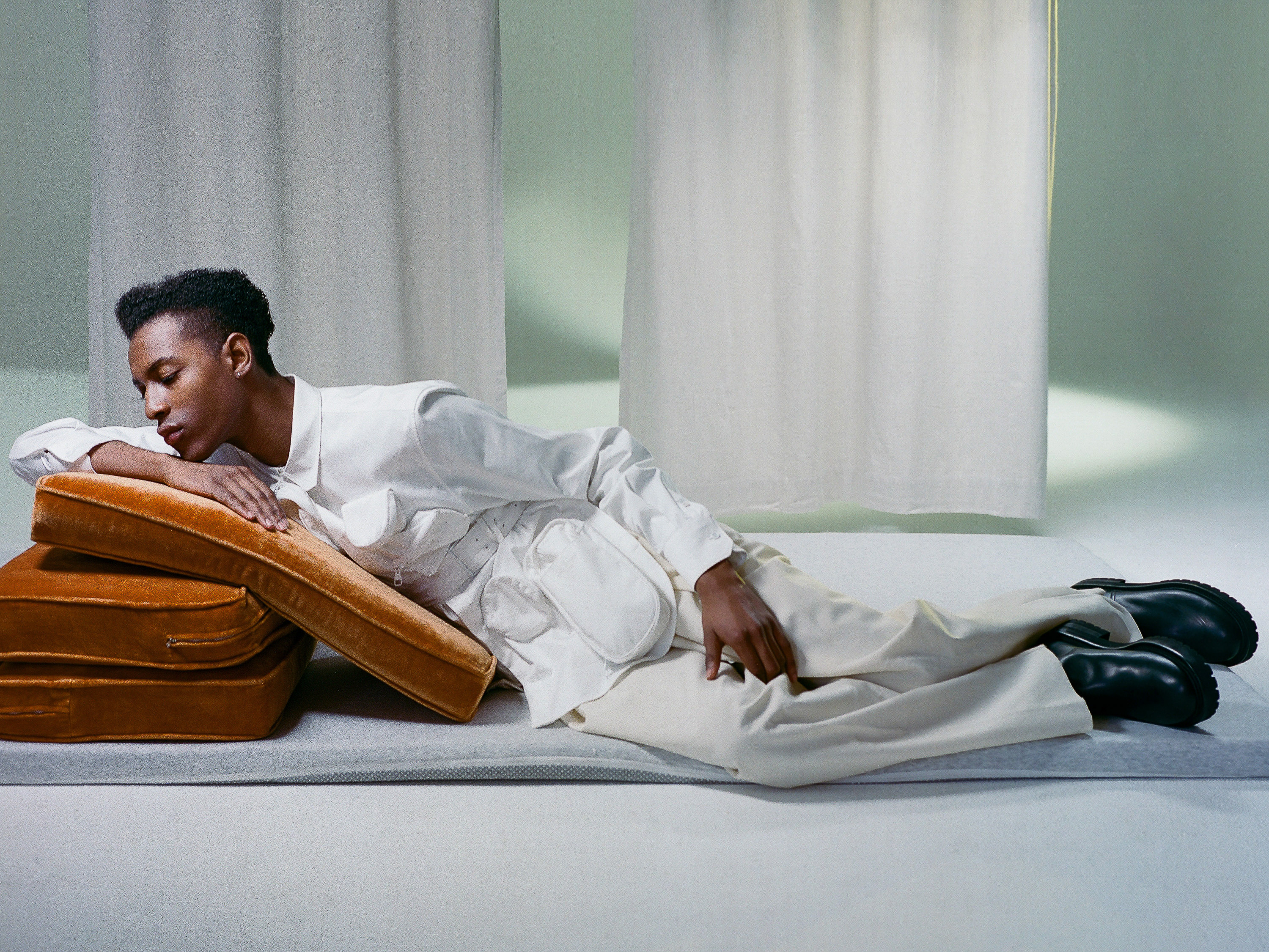 A young adult male model in an all-white outfit rests on brown cushions in a photo studio setting, with white curtains in the background.
