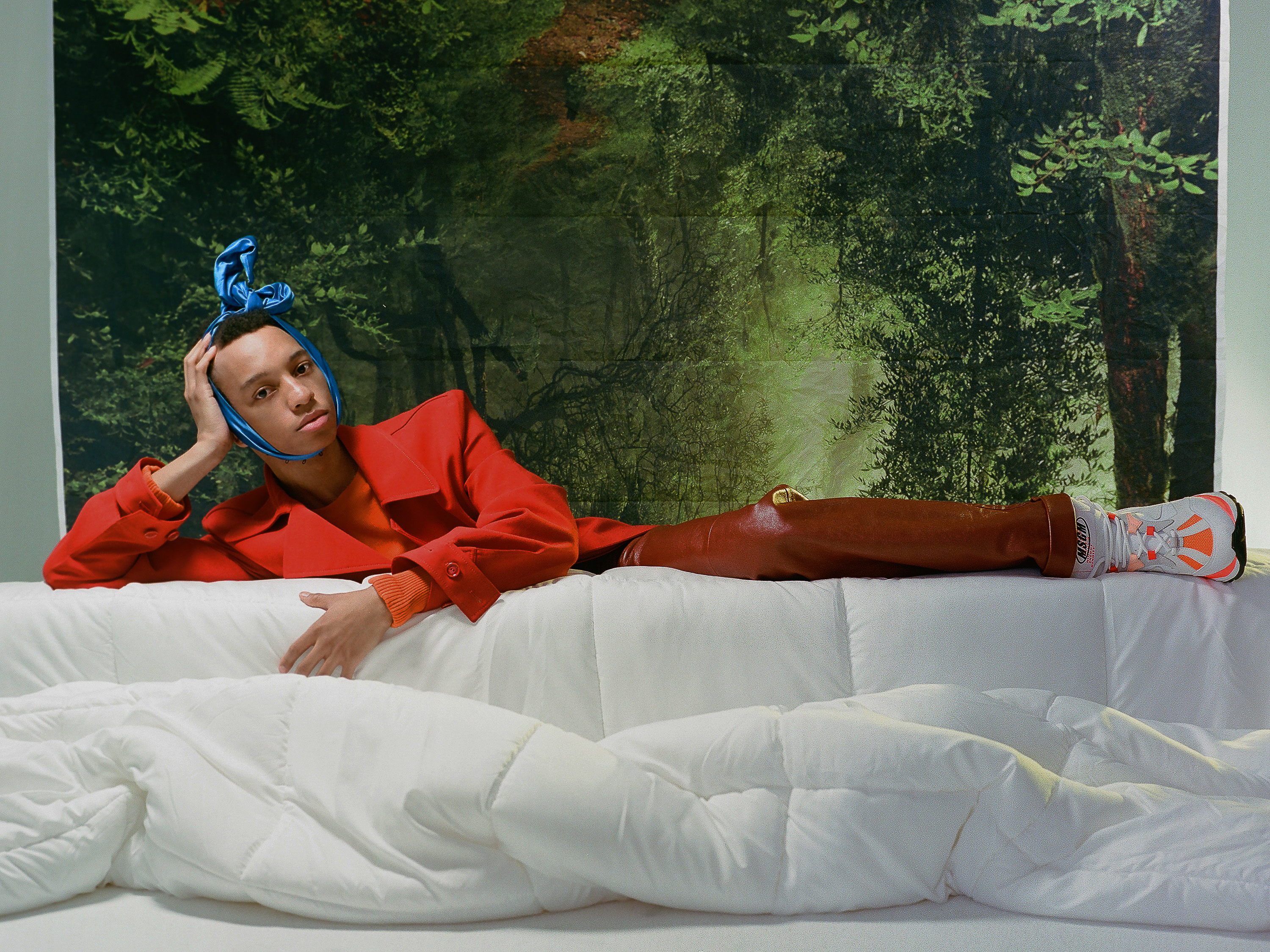 Portrait of a person in a red coat and headscarf reclining on a white sofa against a forest backdrop in a surreal, indoor setting.