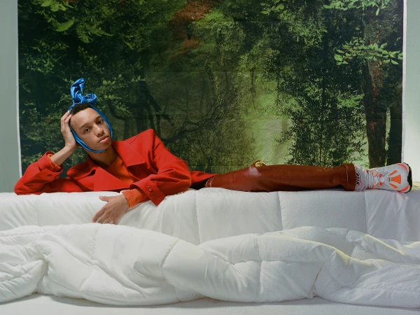 Portrait of a person in a red coat and headscarf reclining on a white sofa against a forest backdrop in a surreal, indoor setting.