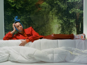 Young man in a red jacket and blue satin head wrap lying on a white quilted bed against a forest backdrop for V Man editorial.