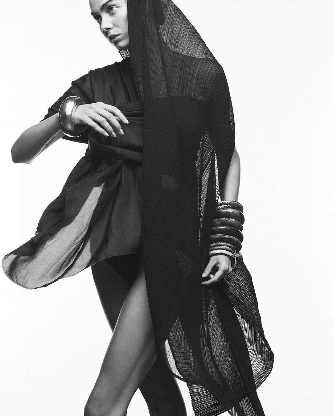 Black-and-white fashion shot of a woman wearing a dark dress, veil, and bracelets, posing against a white background in a studio.