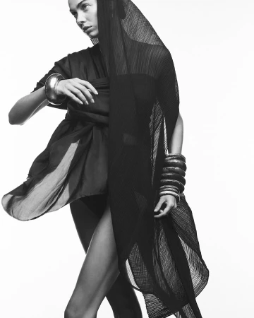 Black-and-white fashion shot of a woman wearing a dark dress, veil, and bracelets, posing against a white background in a studio.