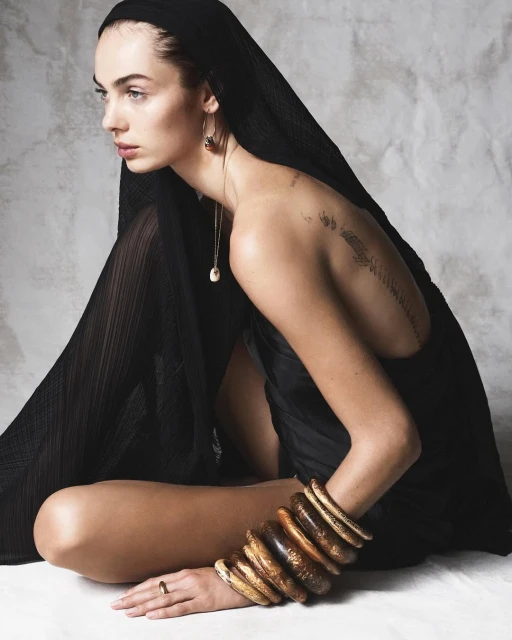 Woman in black headscarf and dress sits with bracelets and a ring against a mottled gray background in a fashion editorial shot.
