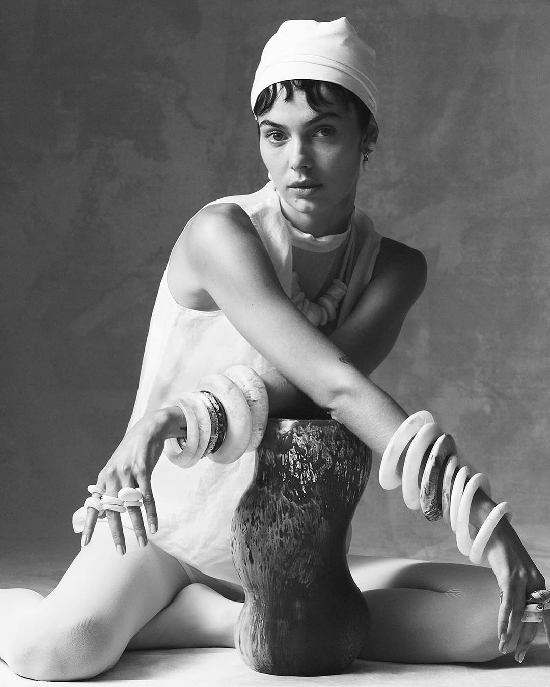 Black and white portrait of a woman in a white headscarf sitting with her arms resting on a vase, wearing large bracelets and a necklace.