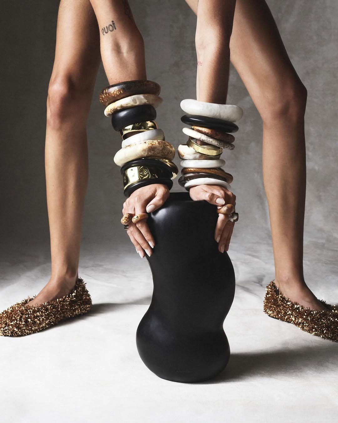 A woman's bare legs and hands adorned with wooden and gold jewelry holding a black vase, featuring golden sparkling shoes on a grey background.