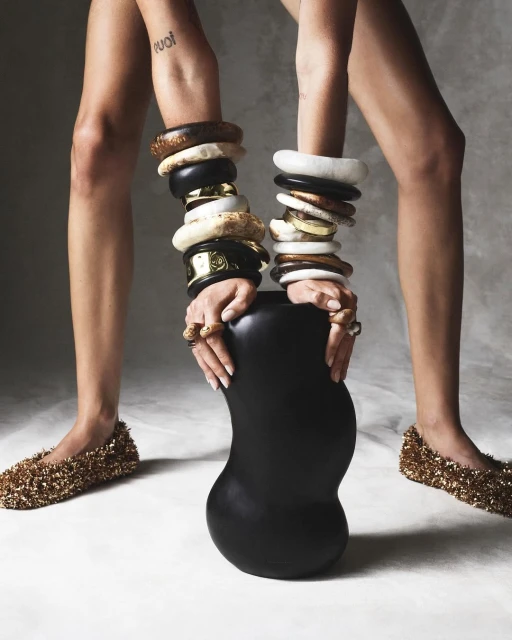 A woman's bare legs and hands adorned with wooden and gold jewelry holding a black vase, featuring golden sparkling shoes on a grey background.