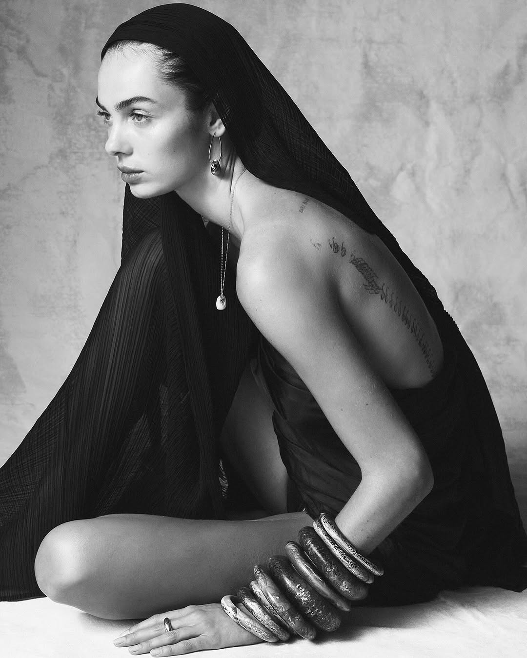 A woman with a black headscarf poses seated wearing bracelets and a ring in a black-and-white fashion photograph with a neutral backdrop.