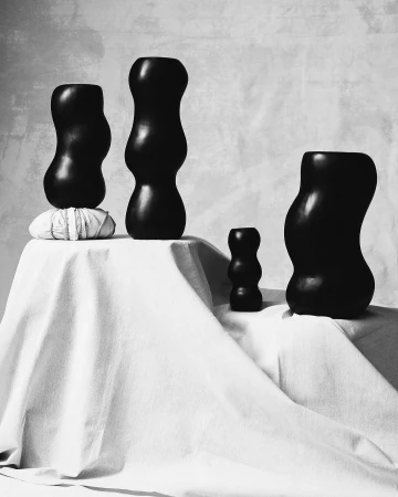 Black vases of varying sizes are displayed on a white fabric-covered surface against a pale grey mottled background, indoors.