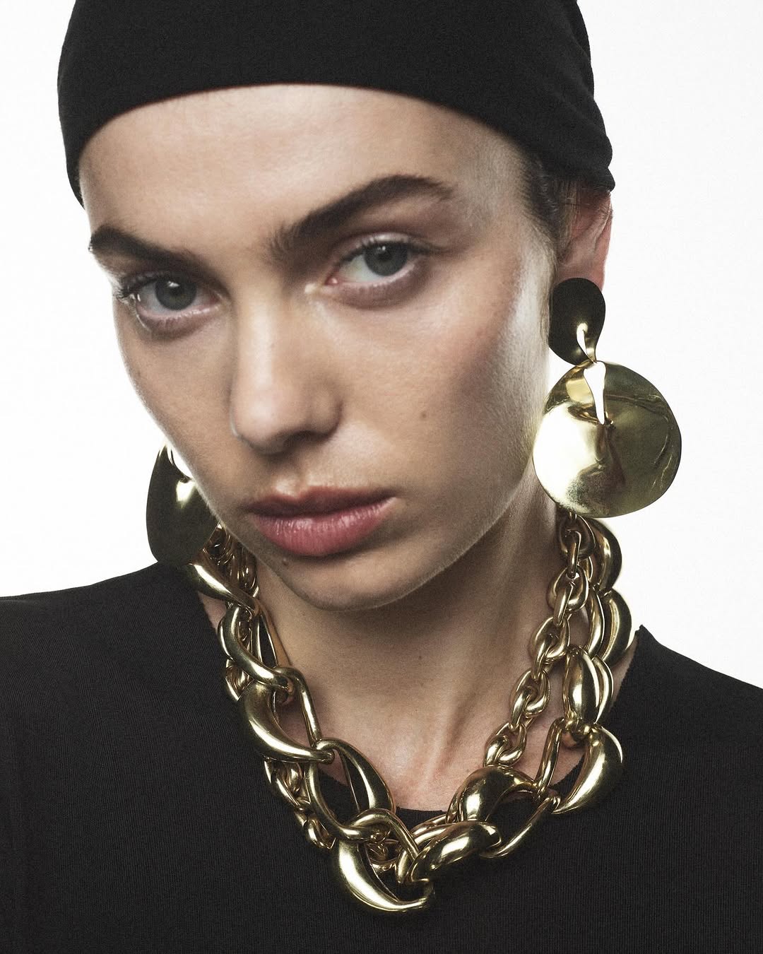 Close-up fashion portrait of a woman wearing a black headscarf, chunky gold necklace, and matching earrings against a white background.
