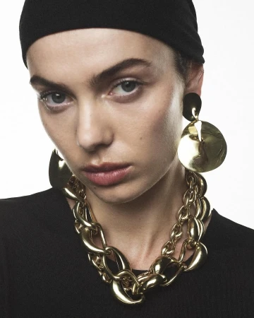 Close-up fashion portrait of a woman wearing a black headscarf, chunky gold necklace, and matching earrings against a white background.