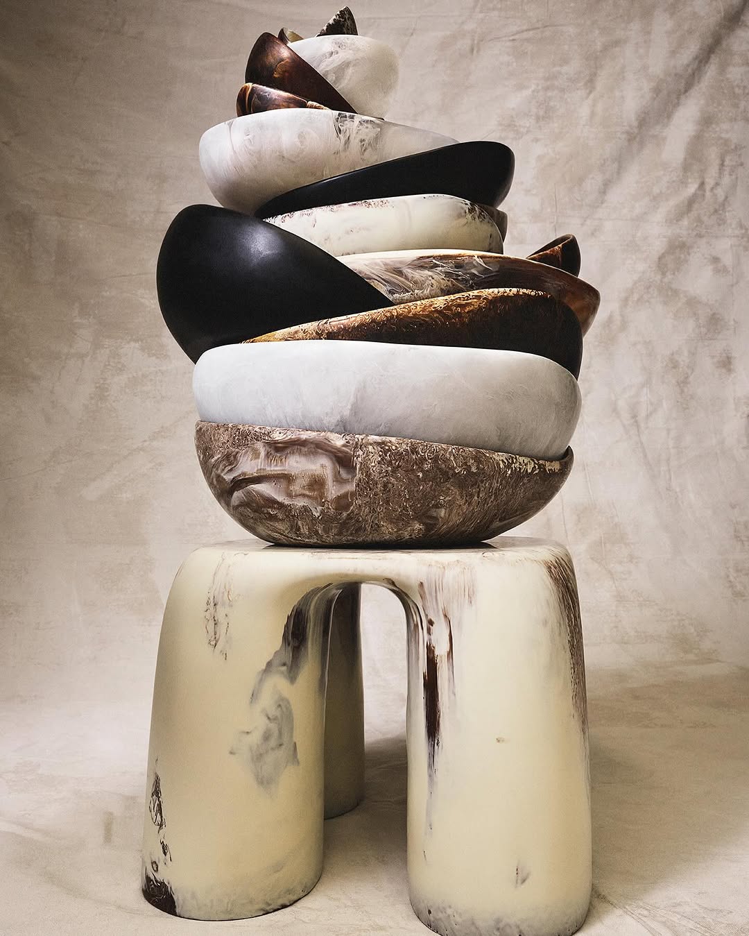 Stacked bowls in various colors and materials sit atop a white stool against a neutral backdrop, creating an artistic still life.