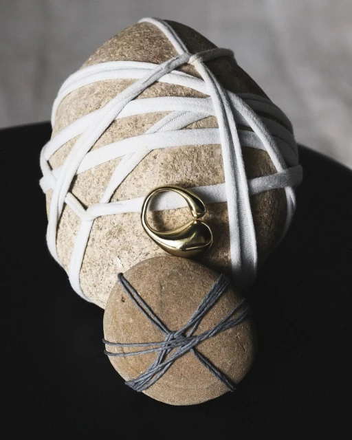 Still life of two rounded stones, each wrapped in different color string, with a gold ring placed between them, against a dark background.