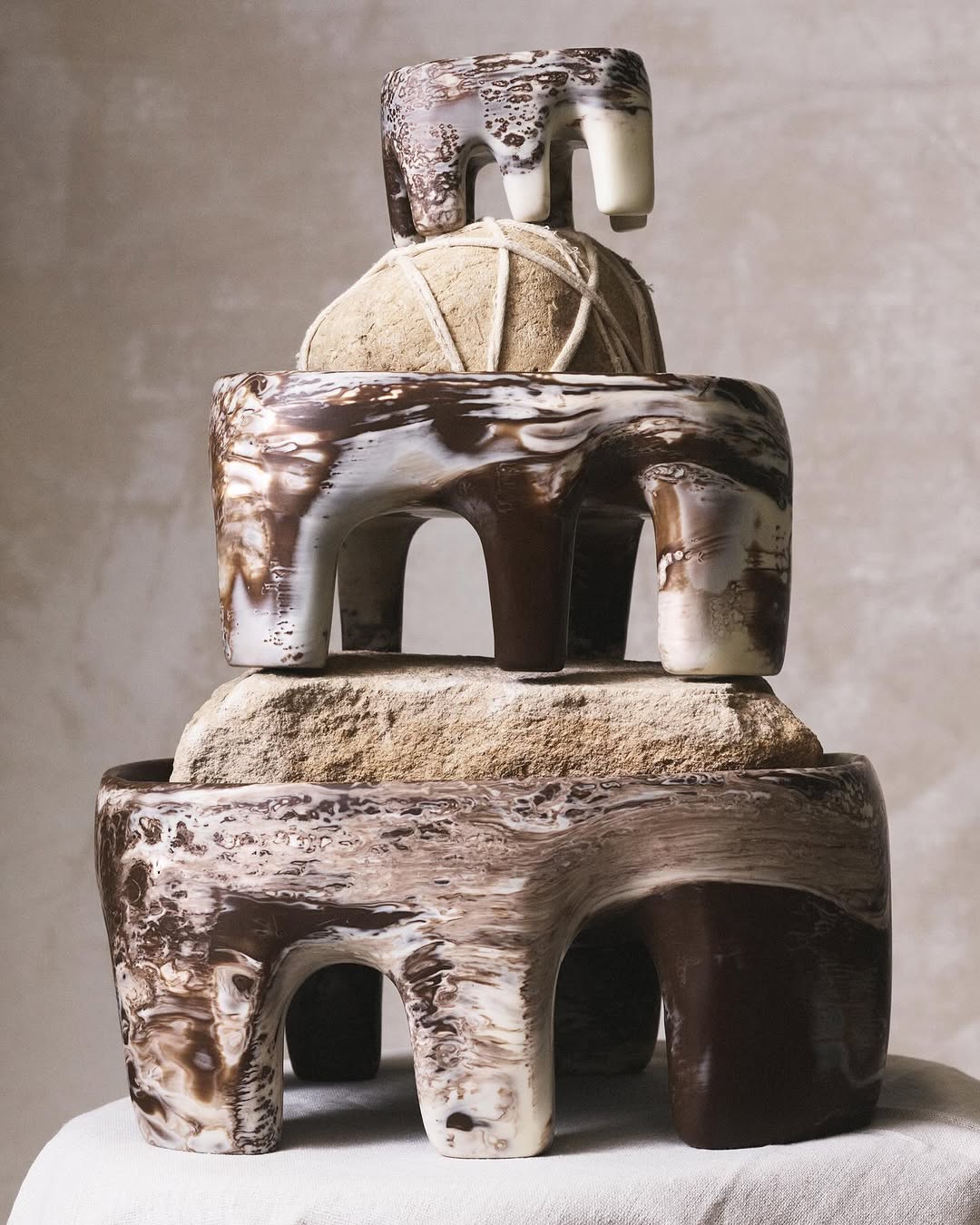 Abstract stack of three ceramic sculptures with white and brown marbling on a white cloth against a light background in studio lighting.