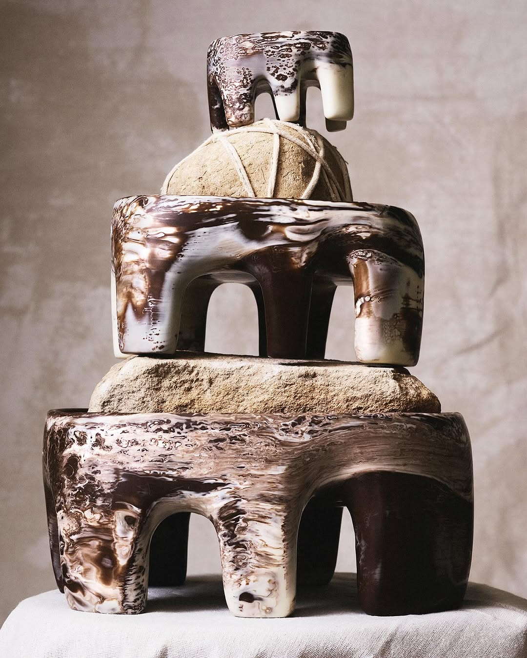 A stack of three abstract sculptures consisting of rounded tan stones and brown and white marbled, four-legged ceramic forms.