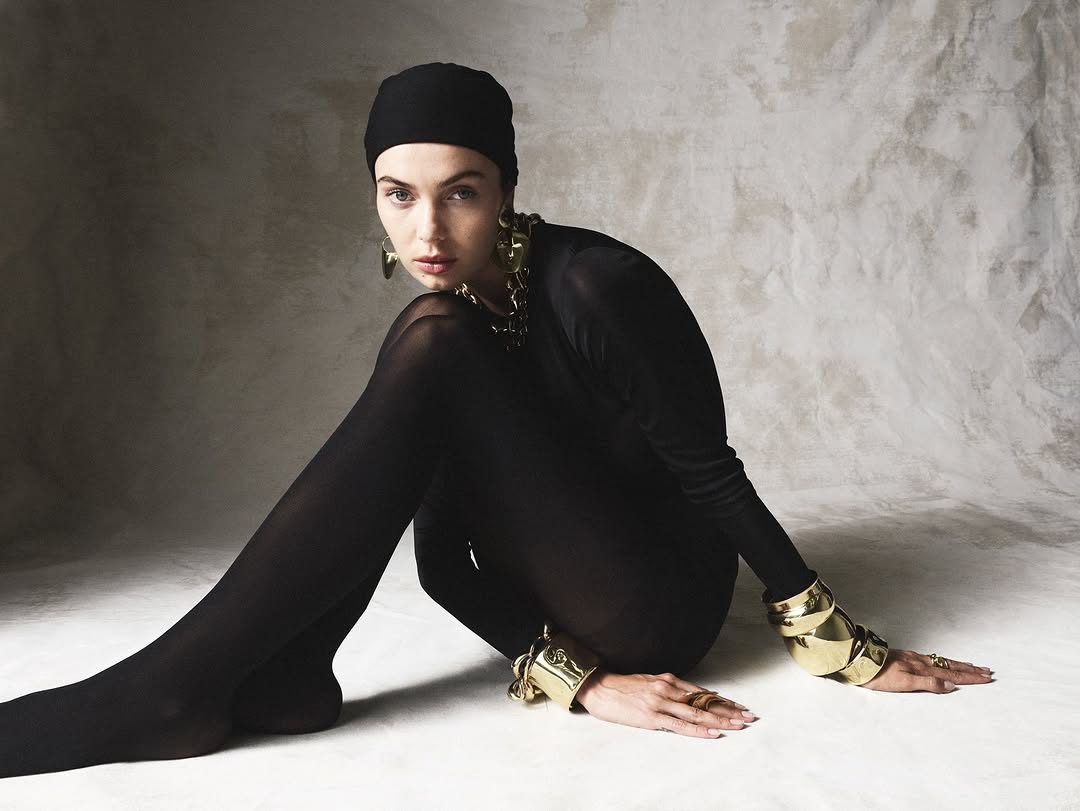 An adult woman poses indoors in a black bodysuit and leggings, accented by gold jewelry, against a neutral textured backdrop.