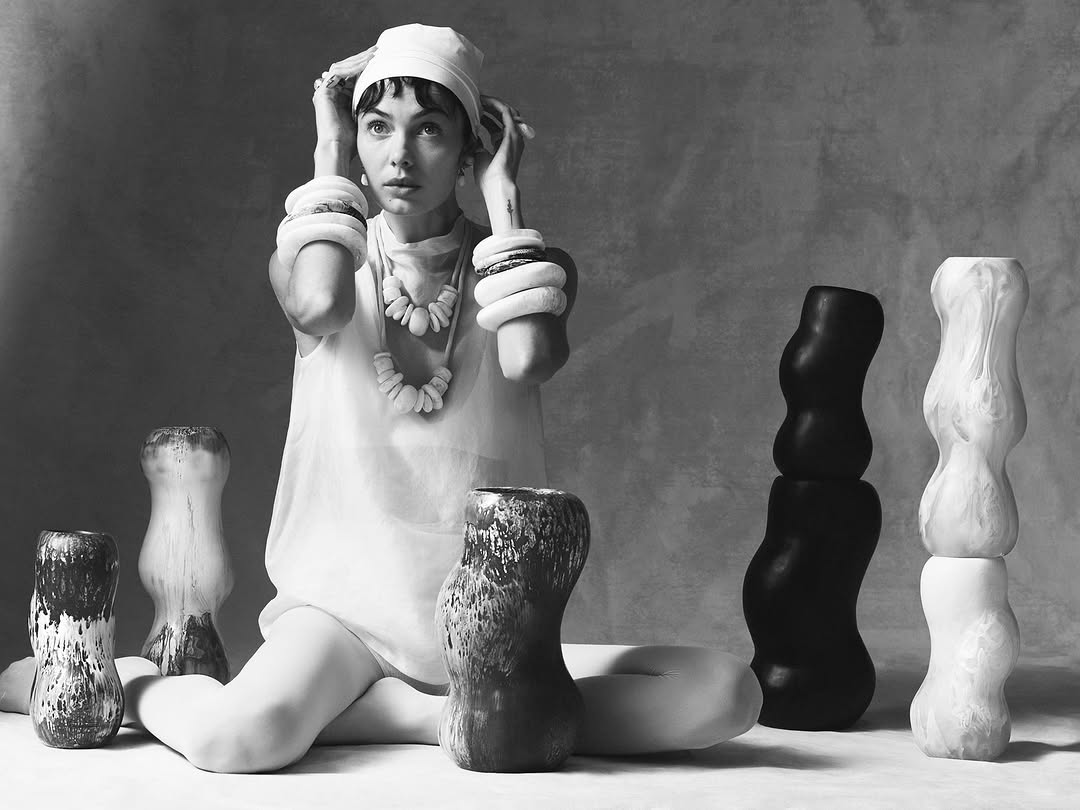 Black and white photo of a woman in a headscarf with bracelets and necklaces, posed in a seated position with vases.