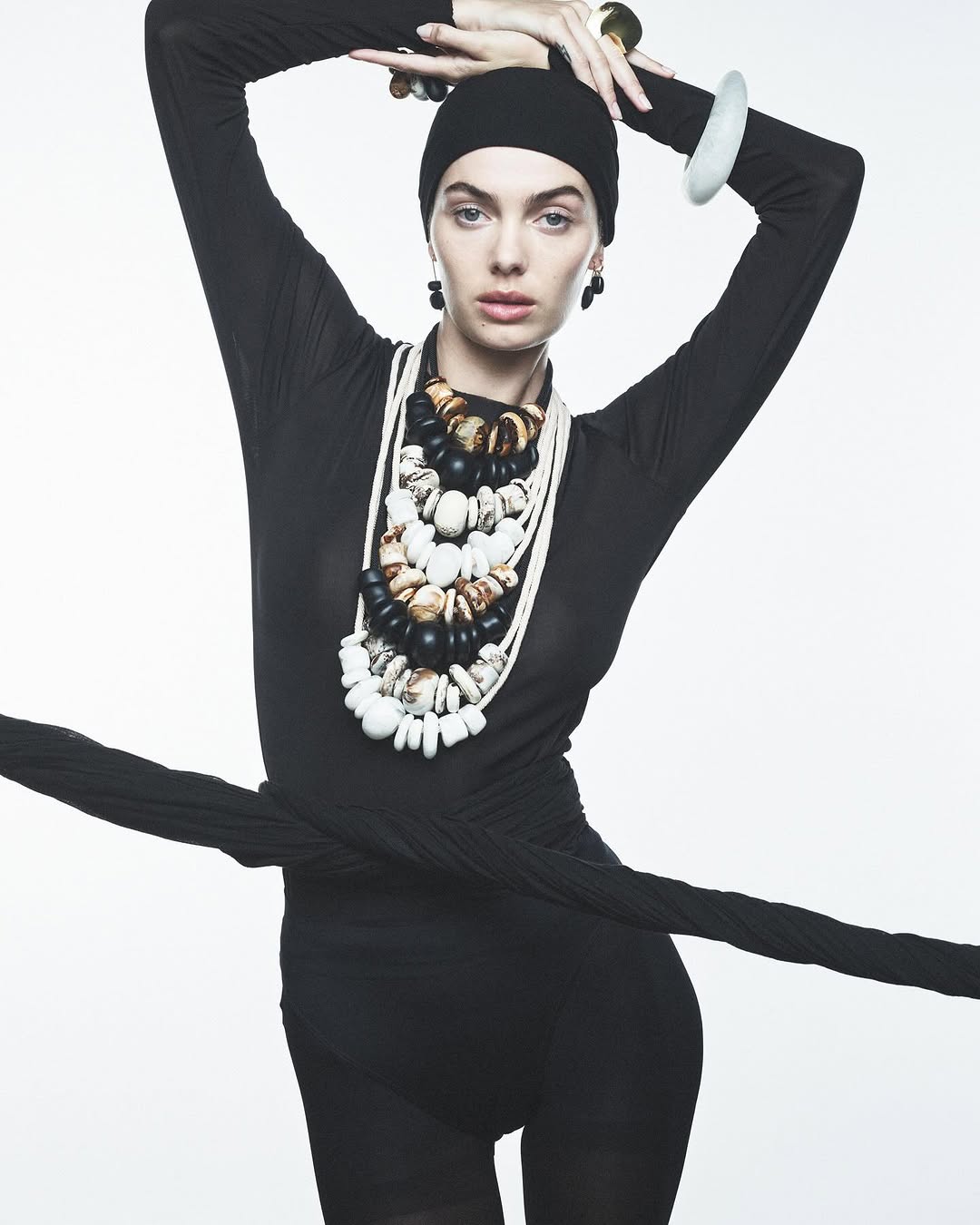 Fashion shot of a woman with beaded necklaces, black head covering, and long-sleeved black garment, set against a white backdrop.