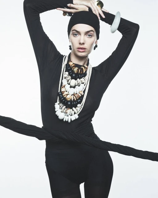 Fashion shot of a woman with beaded necklaces, black head covering, and long-sleeved black garment, set against a white backdrop.