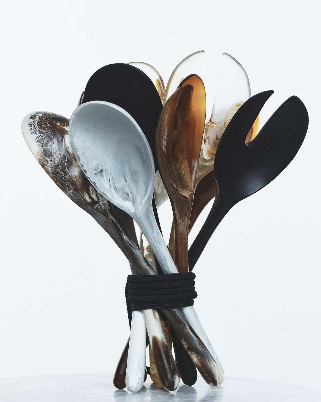Overhead studio shot of black, brown and white salad serving spoons tied together with a black rope against a gradient white background.