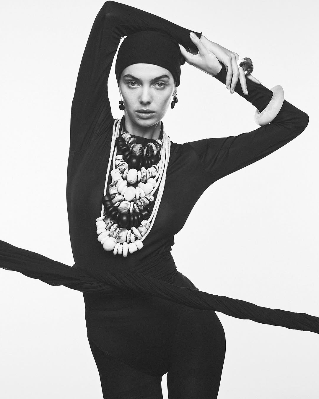 Monochrome fashion shot of a woman in a black bodysuit and head covering, adorned with chunky necklaces and bracelets, against white.