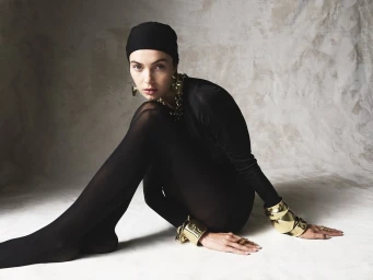 Model wearing black clothing, head wrap, and chunky gold cuffs seated against a textured grey background.