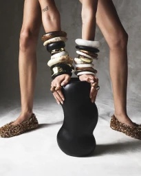 Model wearing stacked chunky bangles and gold textured flats holding a dark sculptural vase