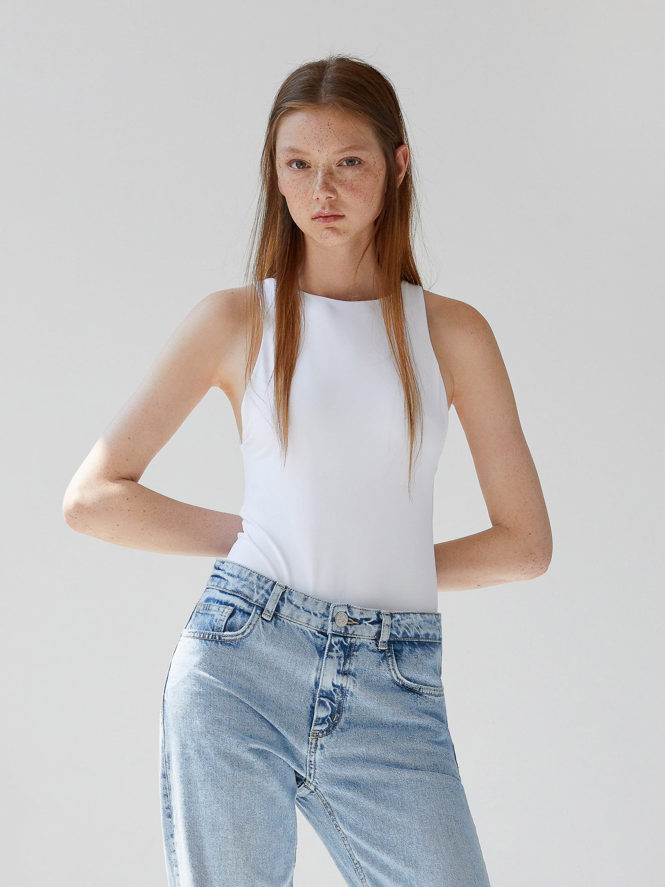 Portrait of a young adult woman with long brown hair posing in a white sleeveless top and blue jeans against a neutral studio background.