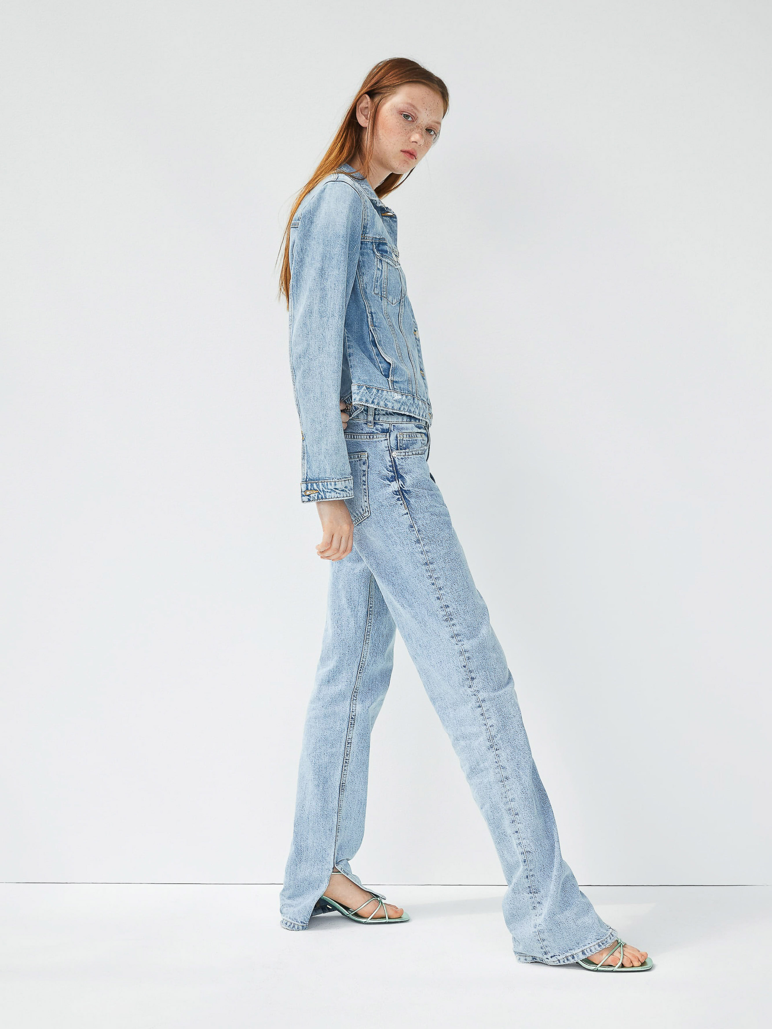 An adult woman with long red hair is standing in a photo studio wearing a light-wash denim jacket and jeans with sandals, against a white backdrop.