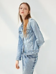 A woman wearing a light blue denim jacket standing against a white wall with soft natural lighting for Zara e-commerce.