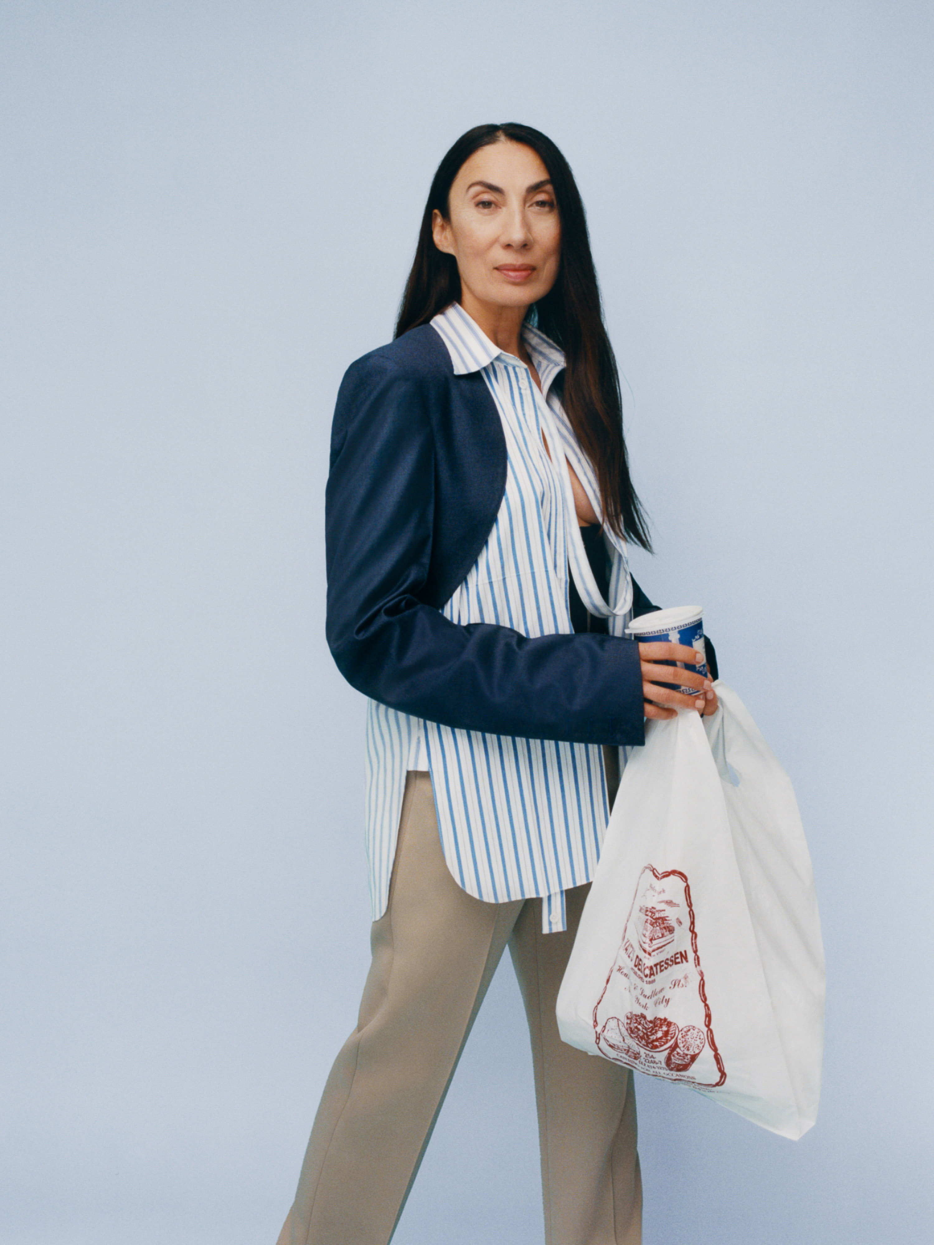Adult woman stands indoors against light blue backdrop, wearing a blazer over a striped shirt, holding a drink and grocery bag.