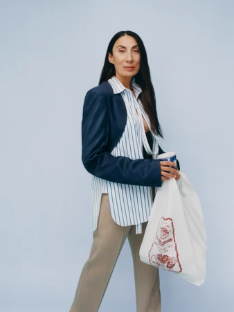 Adult woman stands indoors against light blue backdrop, wearing a blazer over a striped shirt, holding a drink and grocery bag.