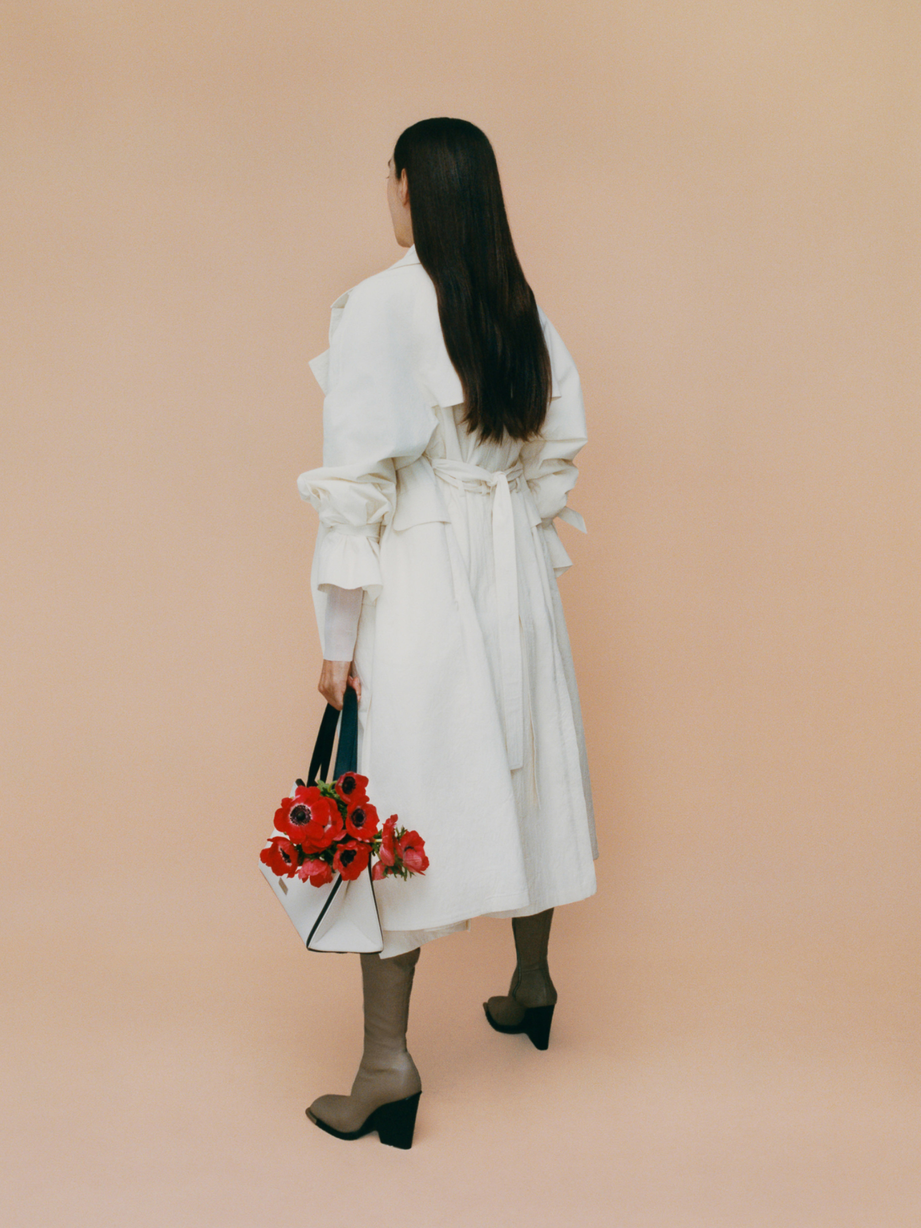 Woman in white coat and brown boots holds a bag of red flowers against a peach backdrop in a fashion editorial shot.