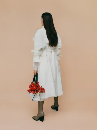 Woman in white coat and brown boots holds a bag of red flowers against a peach backdrop in a fashion editorial shot.
