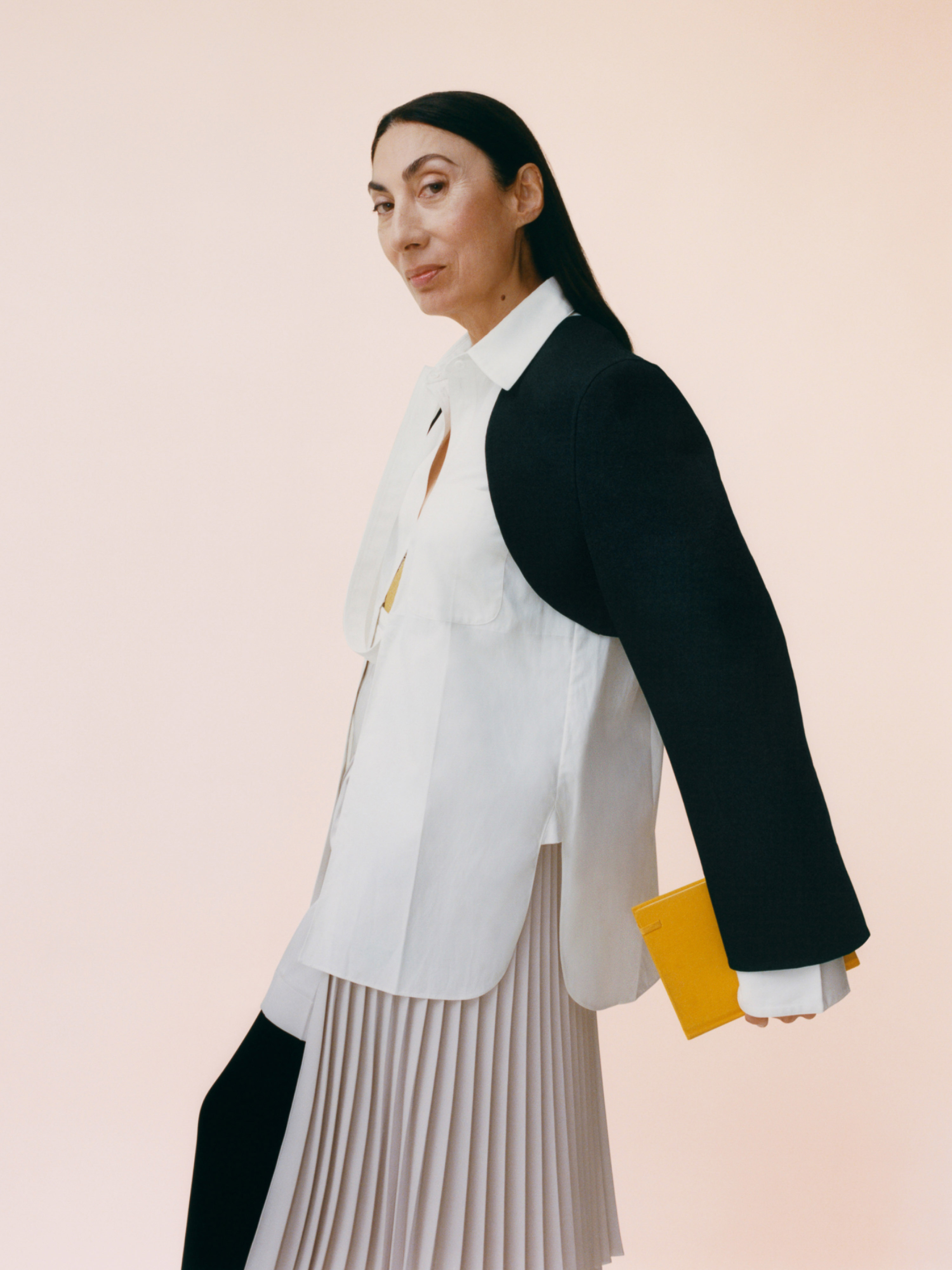 Elegant older woman wearing a white button-down shirt, black jacket detail, and pleated skirt holding a yellow notepad against a peach background.