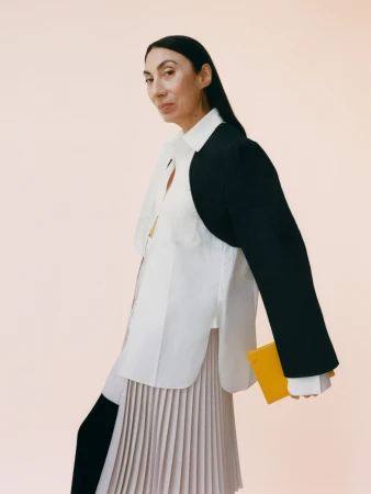 Elegant older woman wearing a white button-down shirt, black jacket detail, and pleated skirt holding a yellow notepad against a peach background.