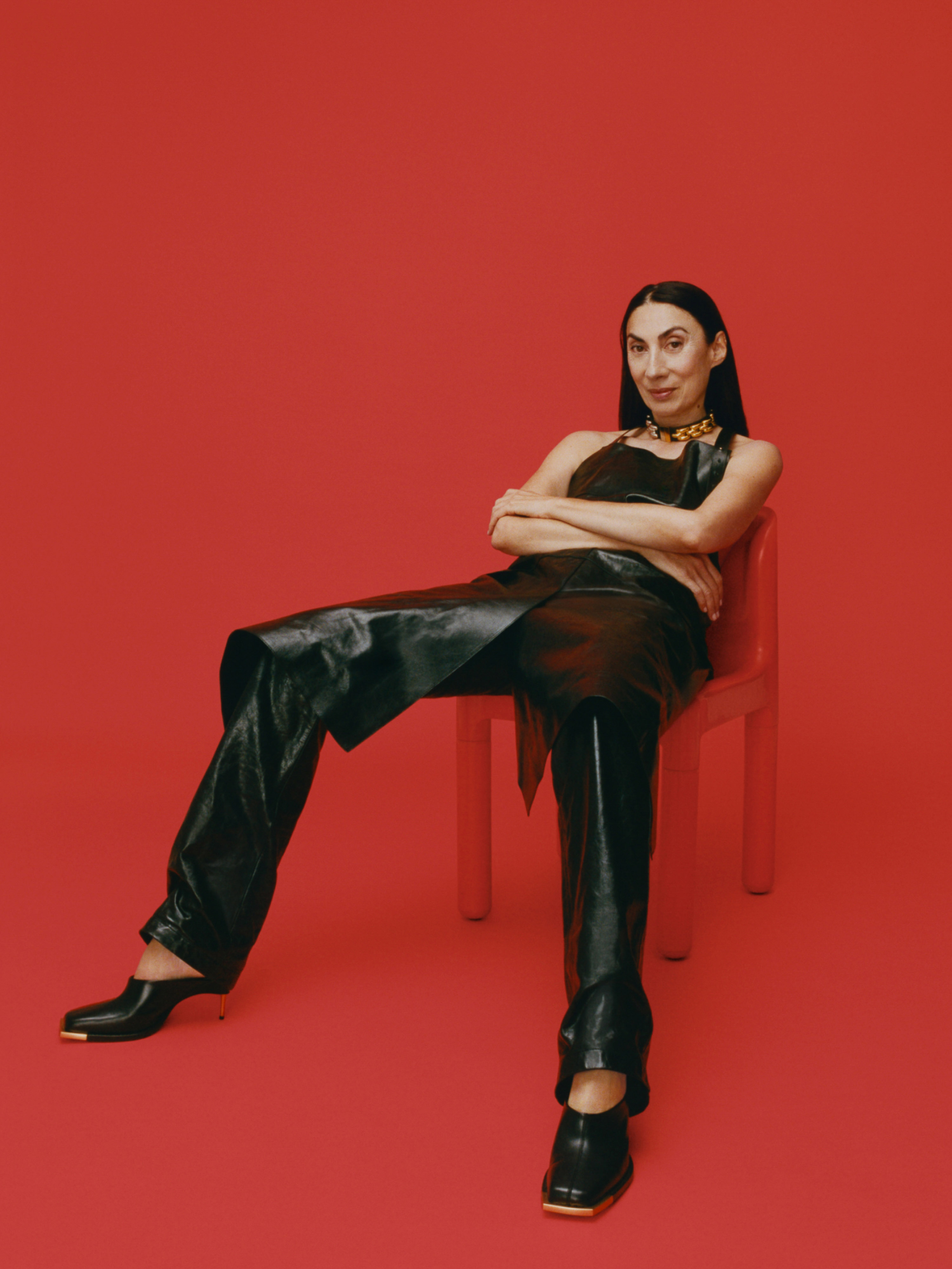 A woman with dark hair in a black top and leather pants sits on a red chair against a red background in a studio setup.