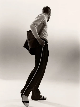 Side shot of a man with a beard in a sweater and track pants, carrying a shoulder bag, against a white background.