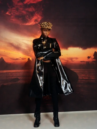 Model in a shiny black patent trench coat and leopard print cap standing before a sunset seascape backdrop.