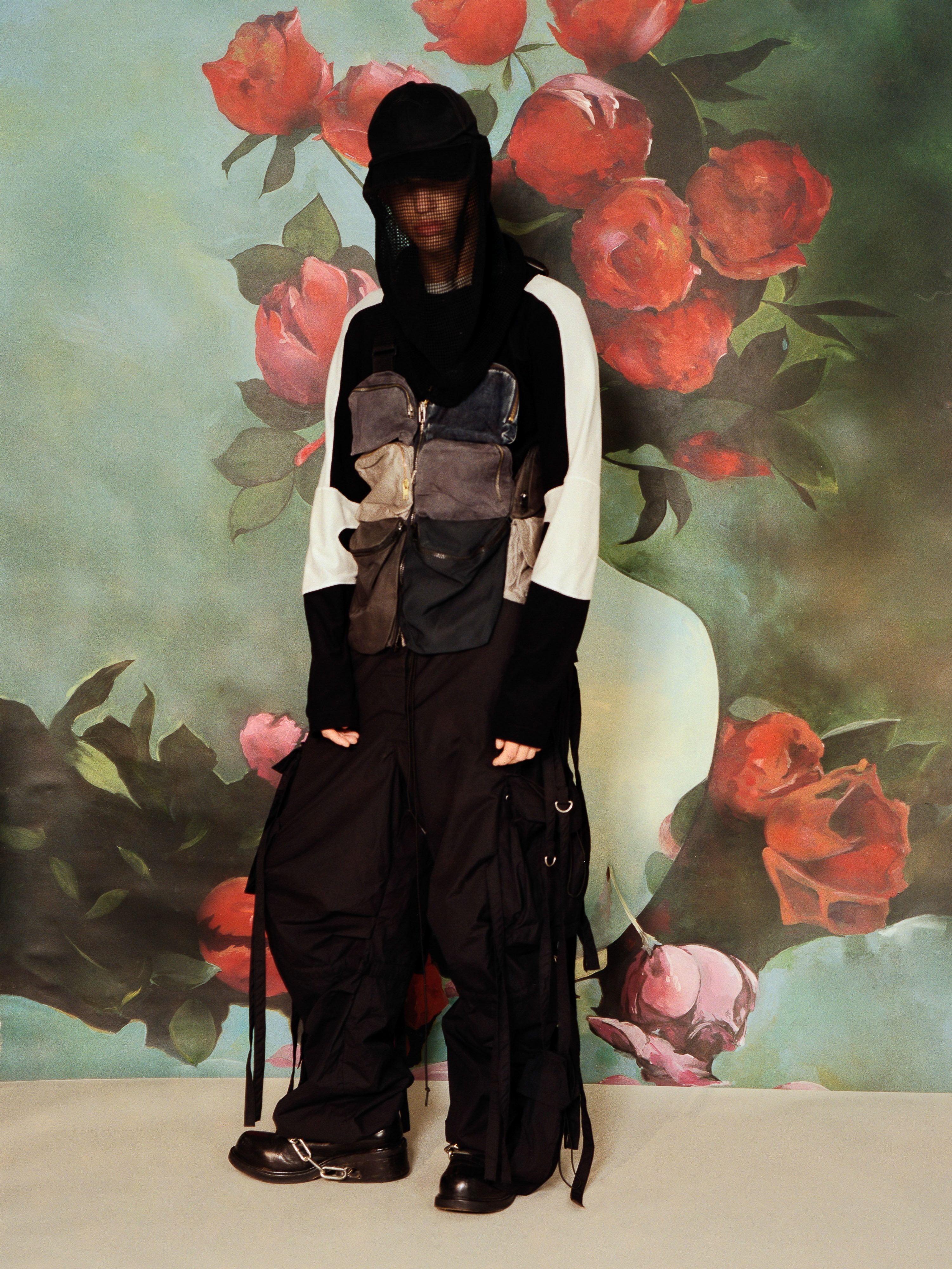 Person in black cap and baggy pants stands in front of rose-covered floral backdrop. Fashion shot with vintage aesthetic and unique clothing.