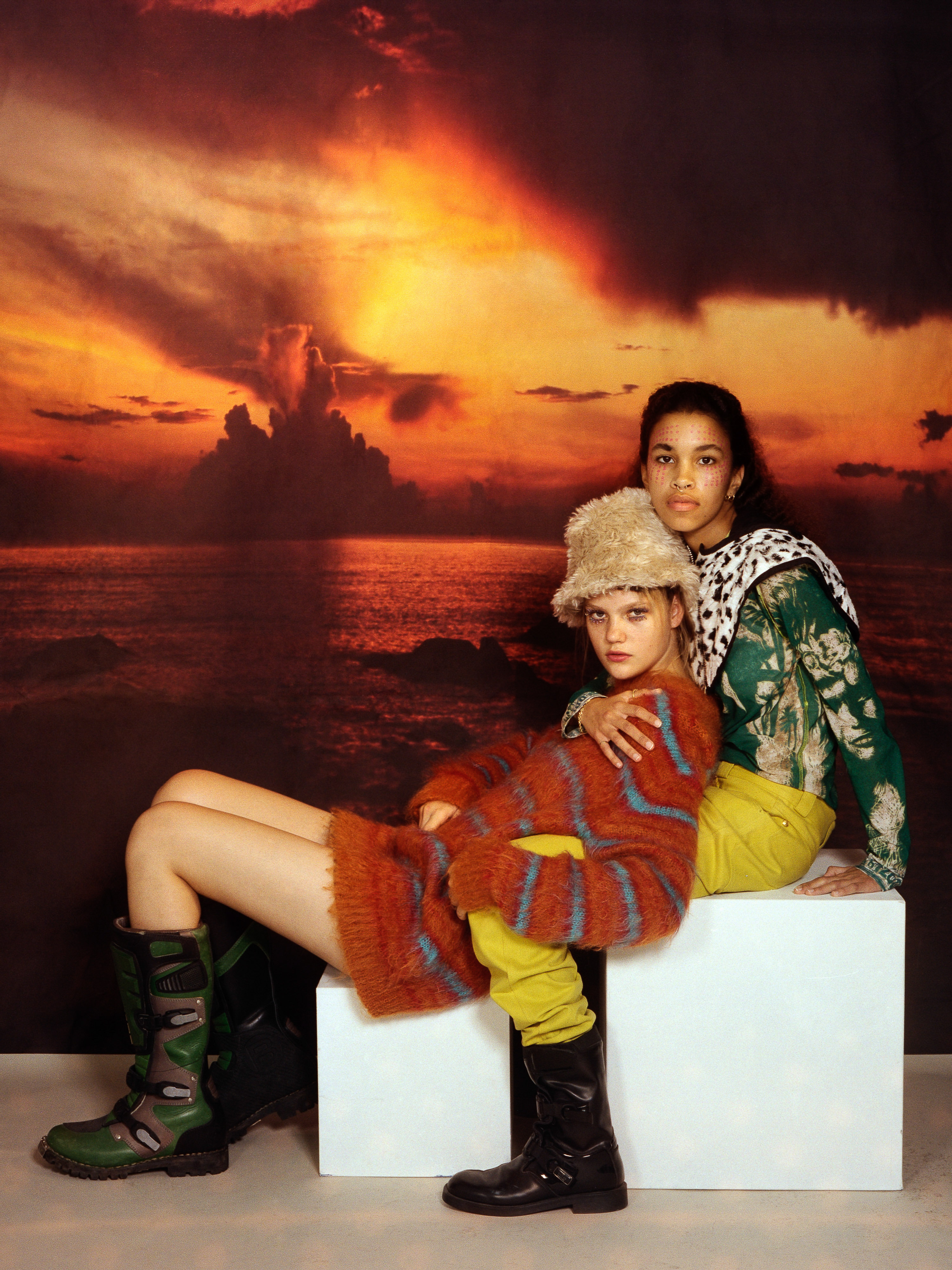 Two young women pose in front of a sunset backdrop, wearing sweaters and stylish boots, creating a fashionable and artistic image.